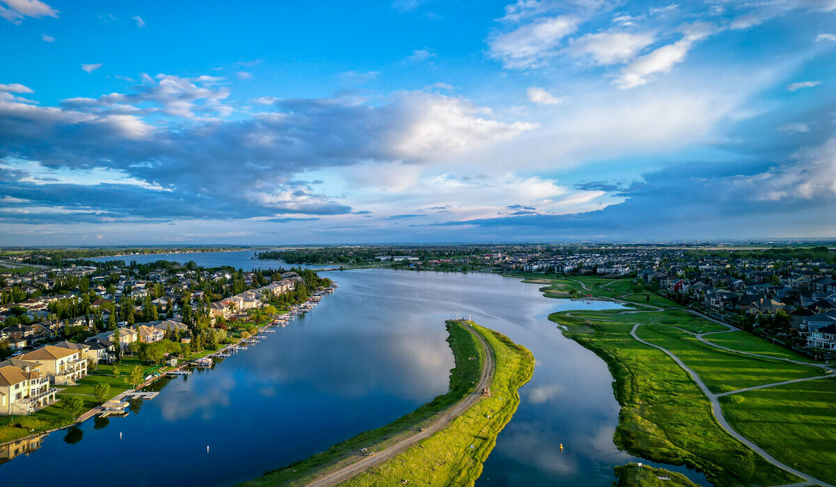 The image is an aerial photo of the City of Chestermere, showing Chestermere Lake with houses lining the shore, surrounded by green areas and roads. The sky is mostly blue with scattered clouds. Across the top of the image is the word "CHESTERMERE" in bold white letters, with the phrase "We want to hear from you!" underneath in smaller white text, set against a blue and green banner.