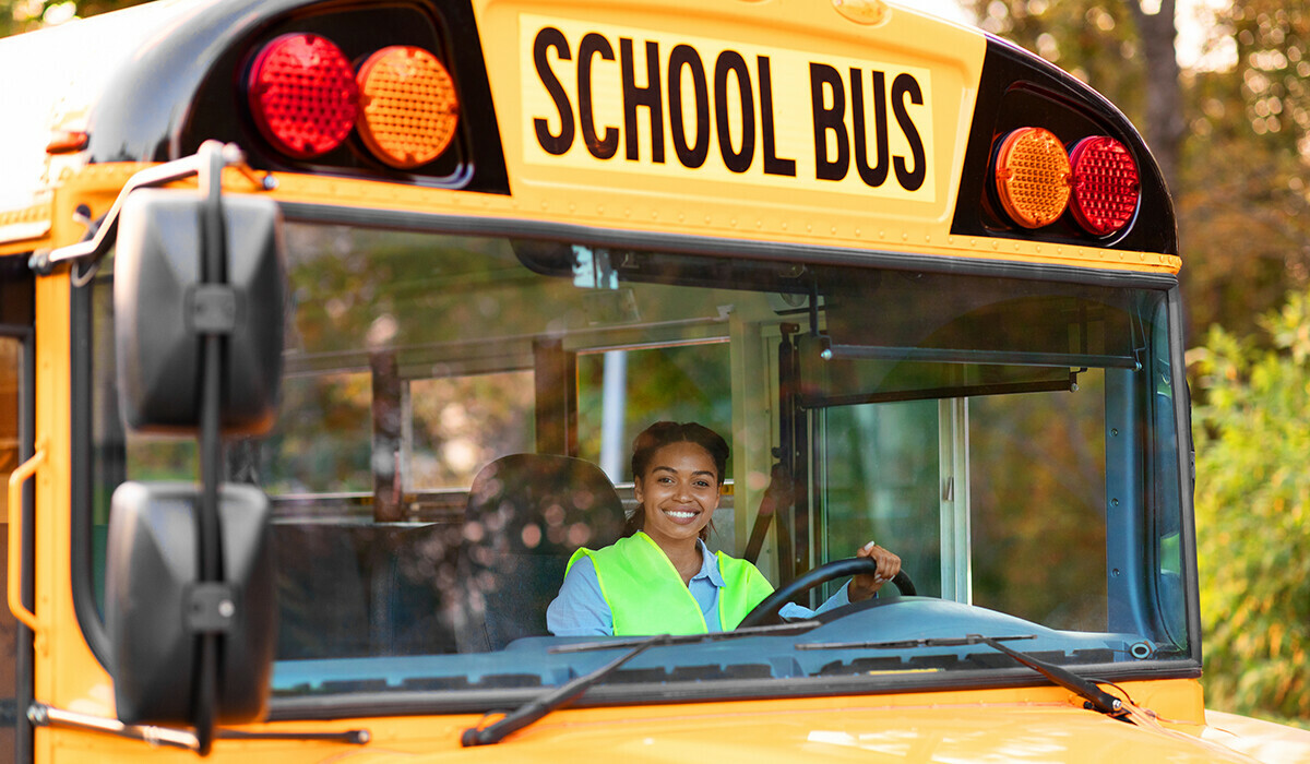 A smiling bus driver wearing a safety vest behind the wheel of a school bus.