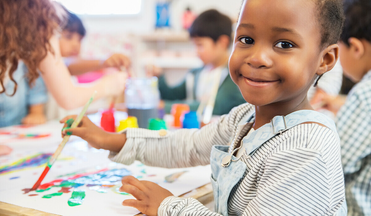 A smiling child in a striped shirt and overalls paints with a paintbrush at a table surrounded by colourful art supplies and other children in a classroom.