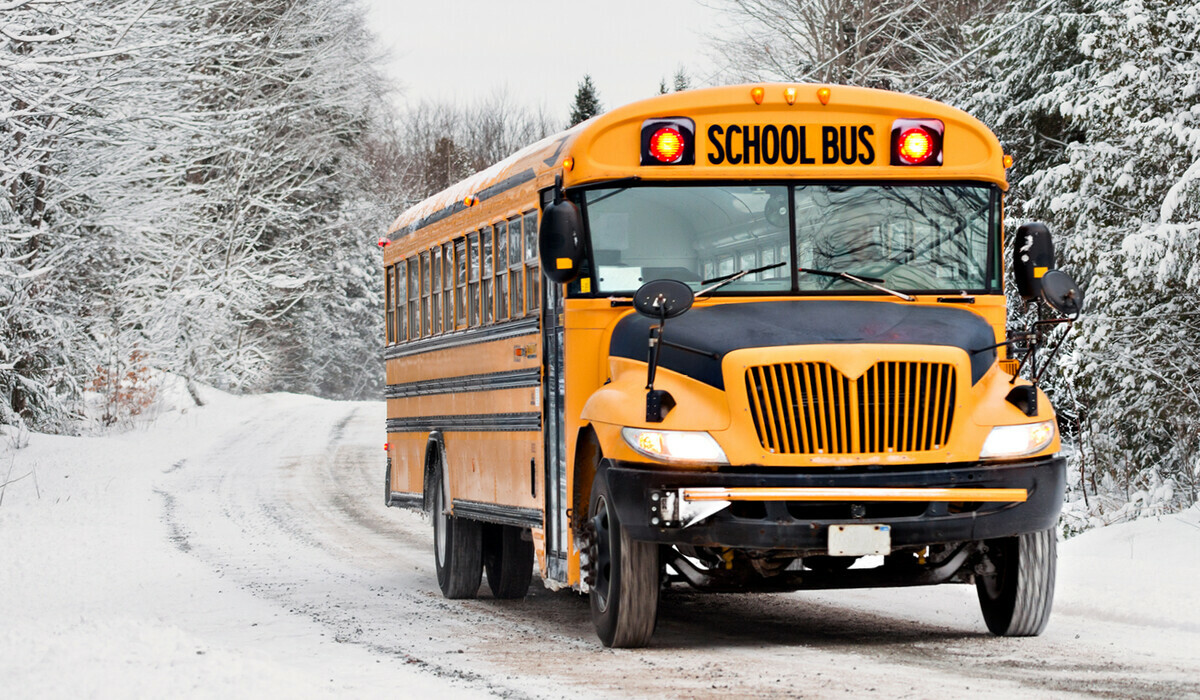 The image shows a school bus driving on a snowy road surrounded by snow-covered trees.